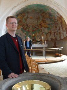 Sognepræst Erik Holmgaard står her ved døbefonten i Gl. Hadserslev Kirke. Både ved dåb og begravelse citerer han teksten fra 1. Peters Brev, som starter med ”Lovet være Gud”. I korbuen bag ham ses Hjalte Skovgaards maleri af den store hvide flok foran Guds trone.